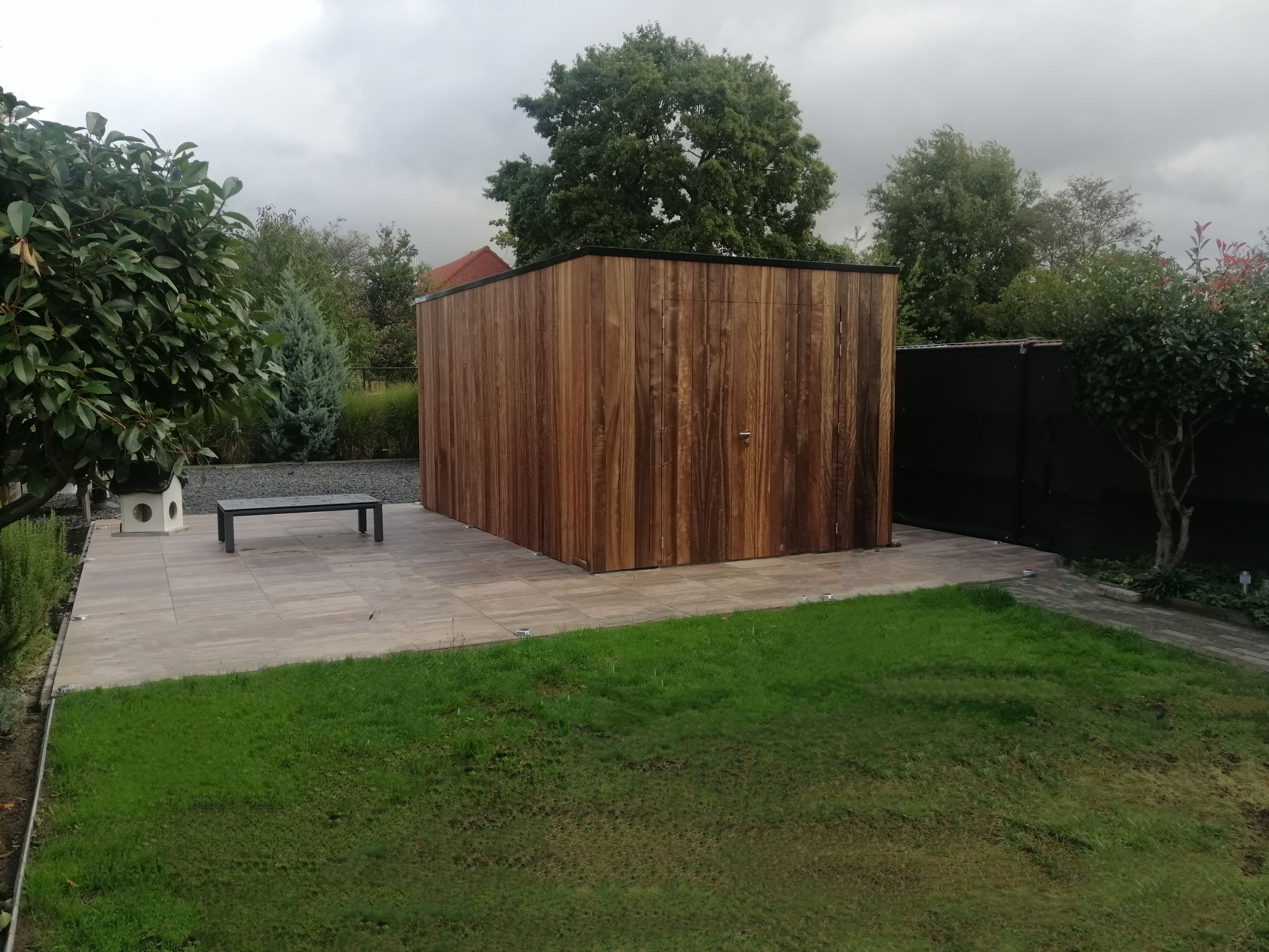 Een houten tuinhuis op betegelde patio met een zwarte bank, omgeven door groen gazon en bomen onder een bewolkte lucht.