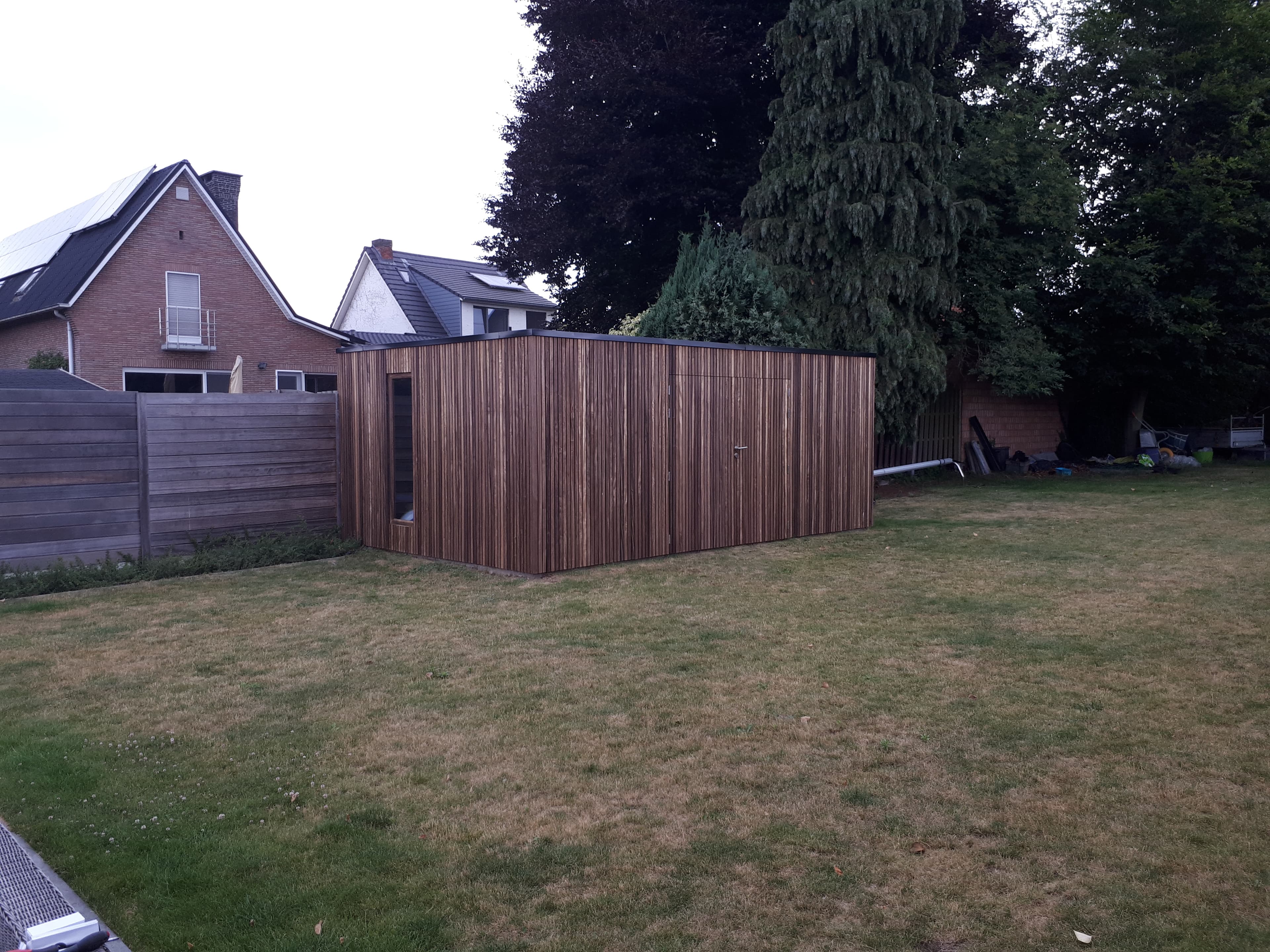 Houten schuur met verticale planken in een achtertuin, omgeven door houten hek en gras, met een grote boom op de achtergrond.