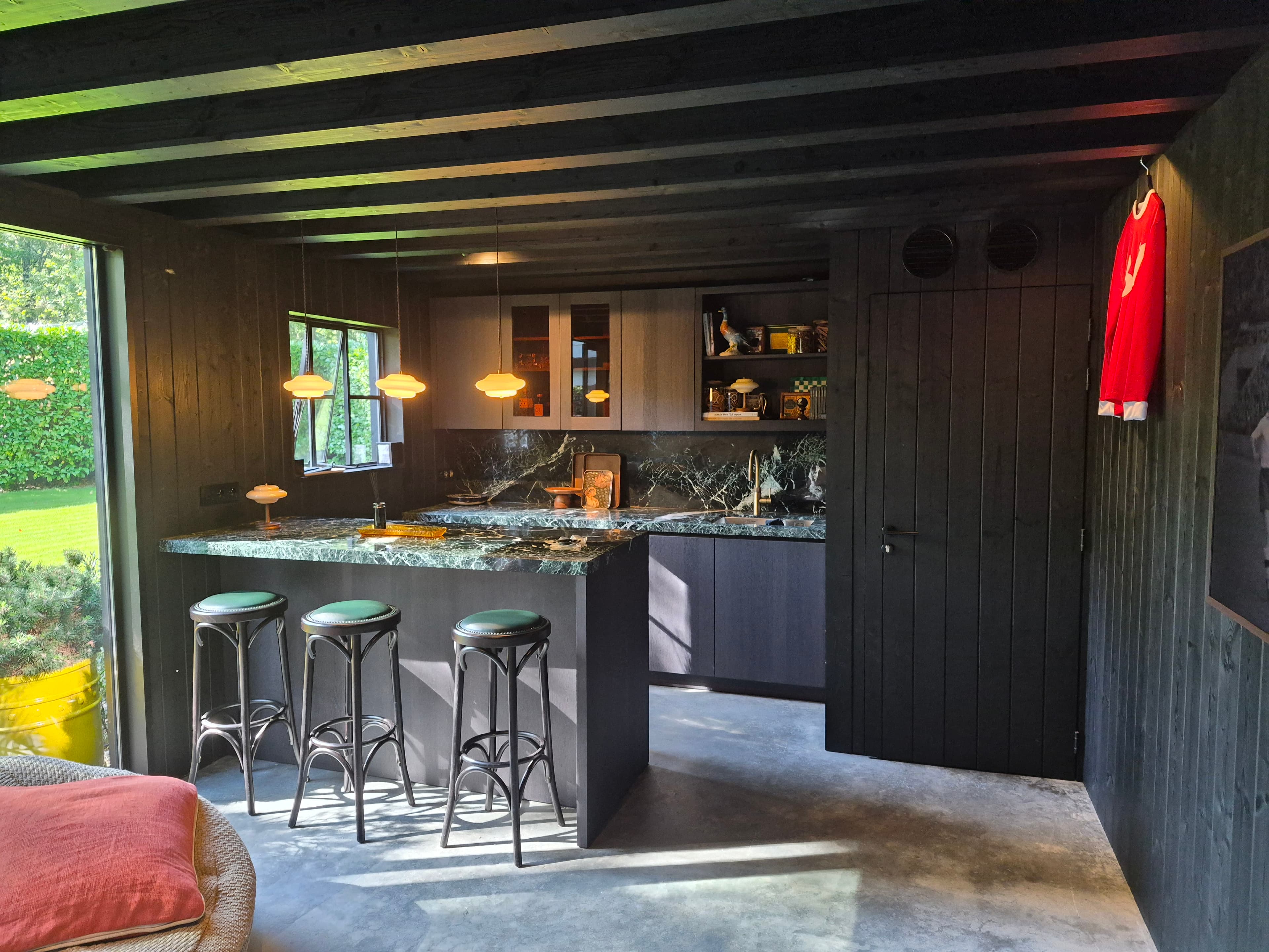 Donkere houten keuken met marmeren aanrecht, barstoelen, hanglampen en een rode jersey aan de muur.