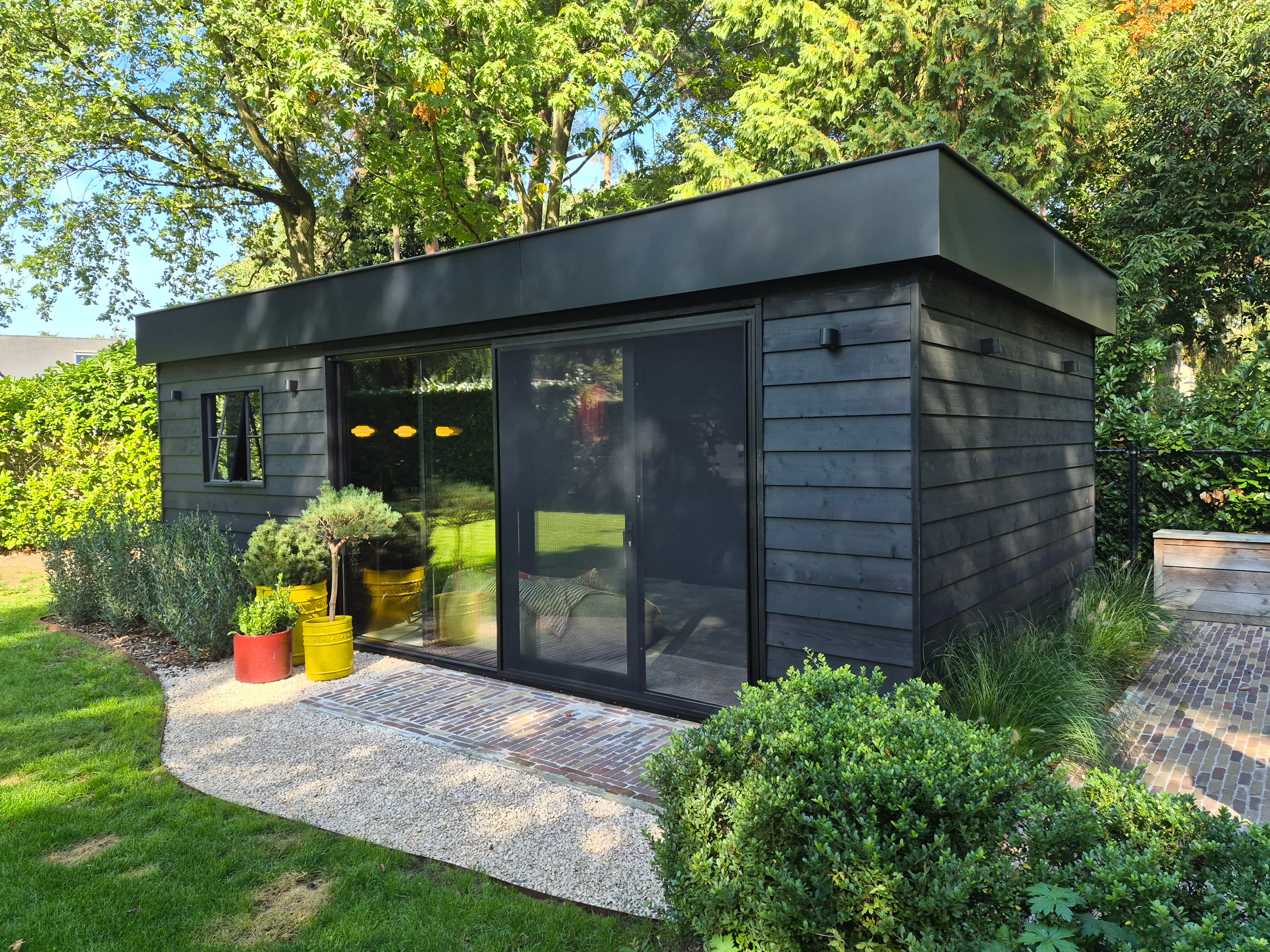 Donkergrijs houten tuinhuis met grote glazen schuifdeur, omgeven door groene bomen en kleurrijke potten langs een grind- en stenen pad.