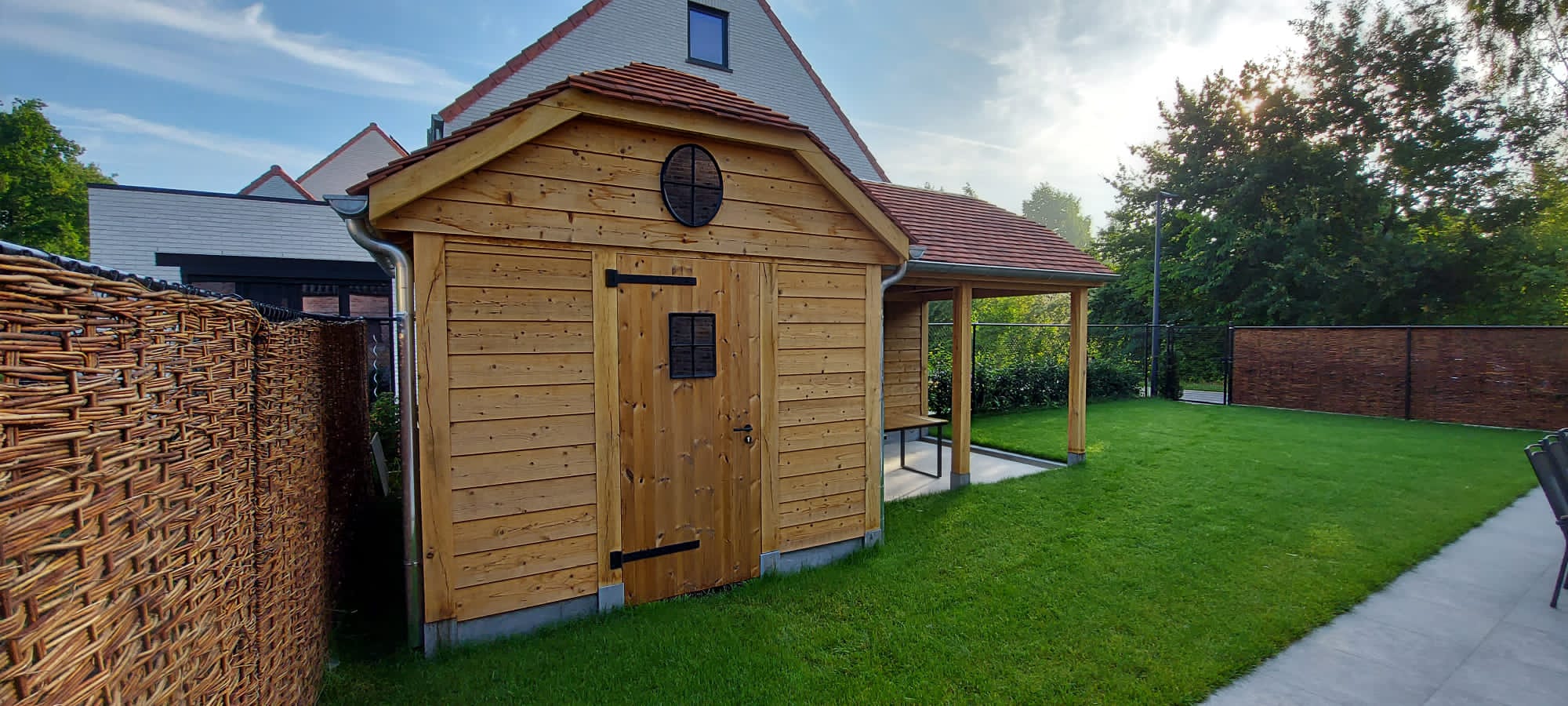 Houten tuinhuis met zadeldak en ronde ventilatie, deur met klein venster, aangrenzende veranda, groene tuin en een riethek aan de zijkant.