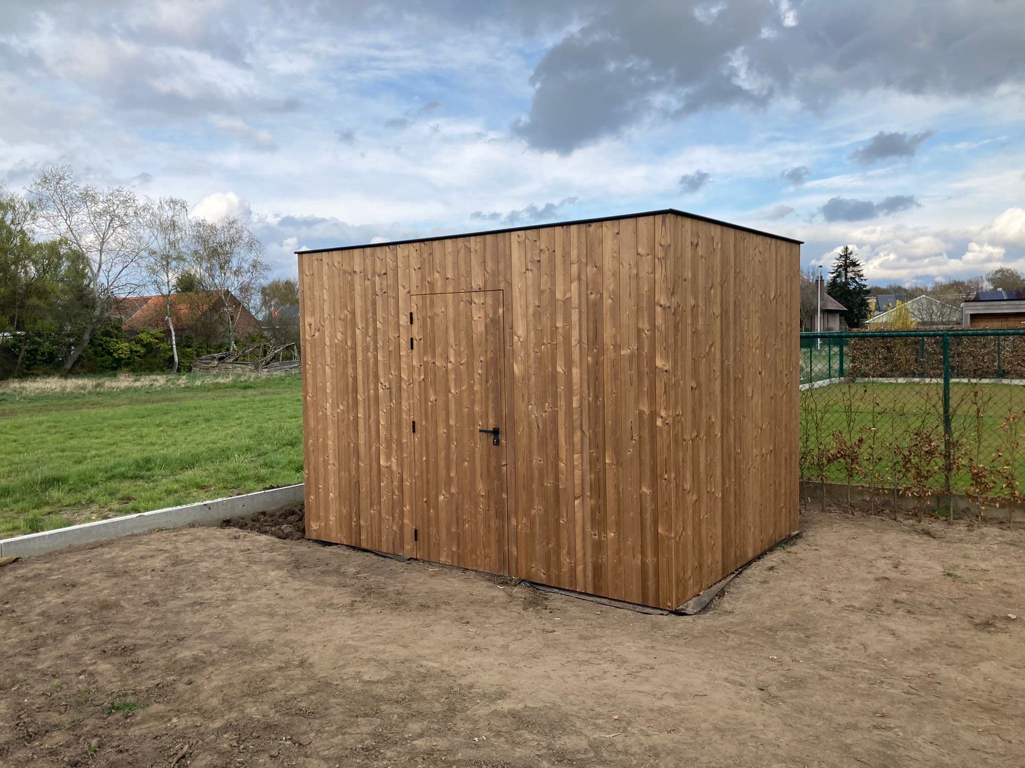 Eenvoudig houten schuurtje in een open tuin, met verticale panelen en een afsluitbare deur, op kaal zand, met bomen op de achtergrond.