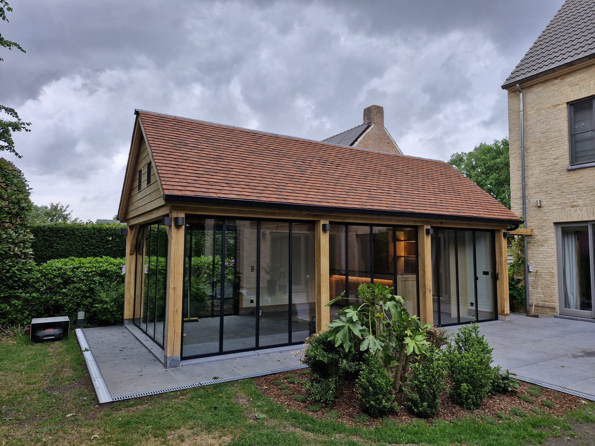 Glazen serre met houten frame en rood dak, naast een stenen huis, in een tuin onder een grijze lucht.