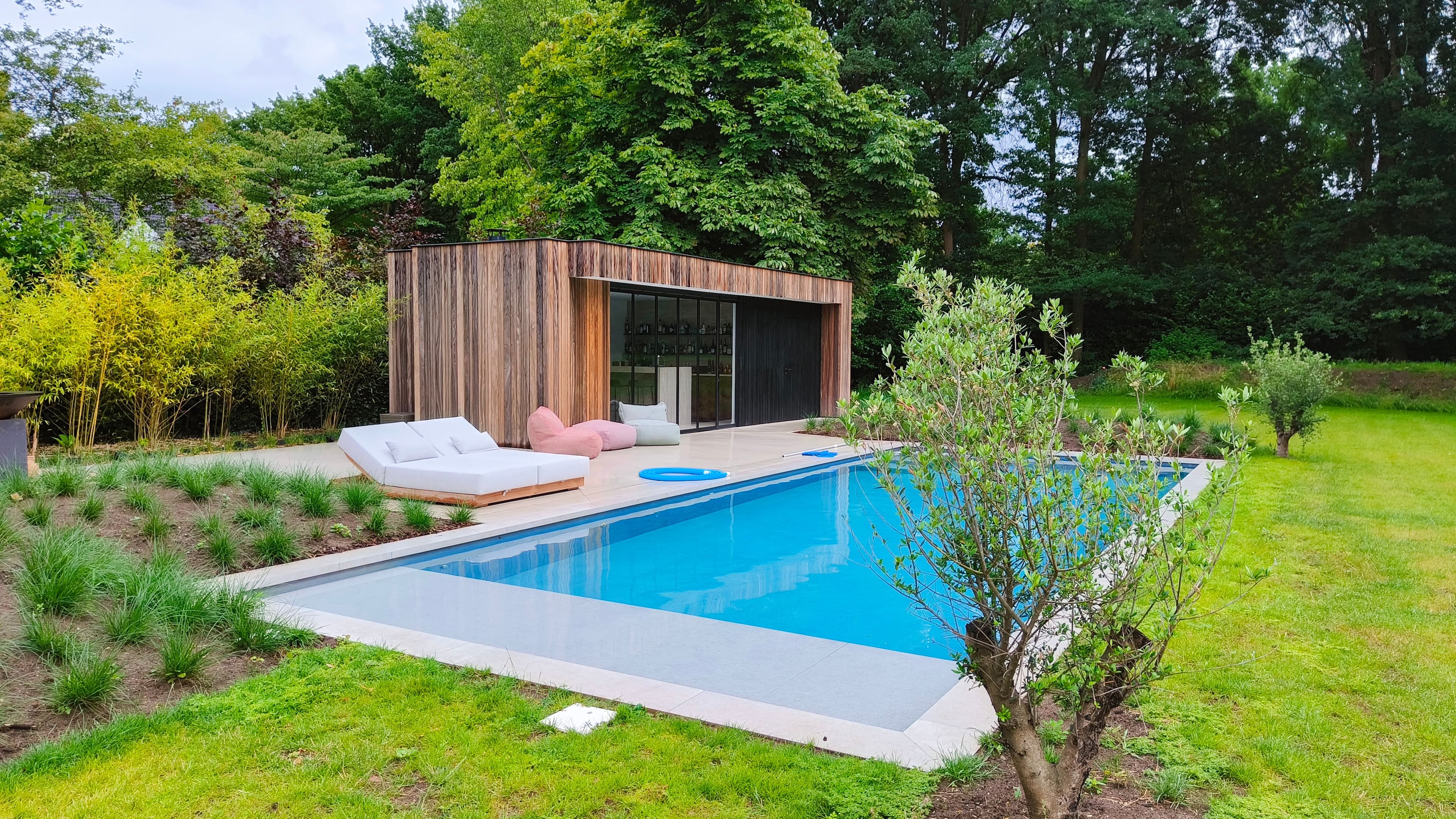 Moderne houten poolhouse op een dek naast een helder blauw zwembad, met witte loungestoelen en roze zitzakken, omgeven door een groene tuin en bomen.